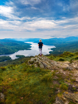 Solo hiker stands on the summit of Ben A'an mountain above Loch Katrine in Scotland, UK, with dramatic clouds, green hills and a calm lake stretching through the Highlands.