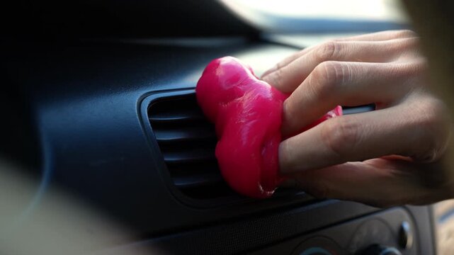 Human hand pressing vibrant pink cleaning gel into grilles of car air conditioning vent, with sticky substance lifting dust and debris from hard to reach areas. Shooting in slow motion.