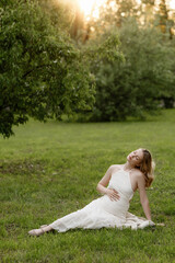 Obraz premium Pregnant woman relaxes on grassy lawn beside lush trees, wearing flowing white dress and leaning back with hand on belly, bathed in warm sunset light, creating peaceful maternity portrait outdoors