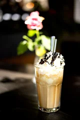 Refreshing iced cappuccino served in a tall glass and topped with whipped cream and chocolate syrup. Soft floral bokeh and warm natural light create a cozy outdoor café atmosphere. © Alejandro