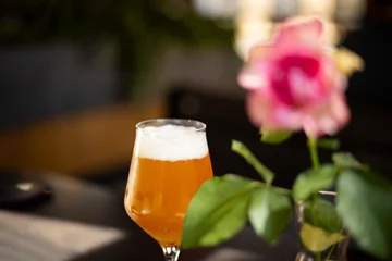 Golden craft beer served in an elegant tulip glass with a creamy foam head at an outdoor café. A softly blurred pink rose and natural bokeh create a relaxed summer atmosphere with selective focus. © Alejandro