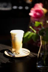Freshly prepared hot cappuccino served in a clear glass with creamy milk foam on a ceramic saucer. Natural sunlight and soft floral bokeh create a warm outdoor café atmosphere. © Alejandro