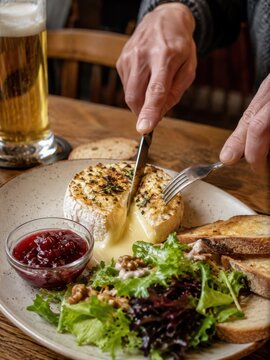 Ofengebackener Camembert wird im Restaurant angeschnitten, der warme K&auml;se l&auml;uft cremig heraus. Serviert mit Preiselbeeren, Salat, Waln&uuml;ssen, Brot und einem Glas Bier.