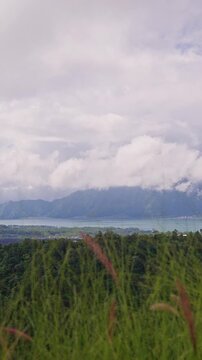 Vertical video. Scenic View of Lake and Mountain Range Beyond Tropical Hills Framed by Tall Grass and Rolling Clouds. Travel Destination, Natural Beauty, Serenity