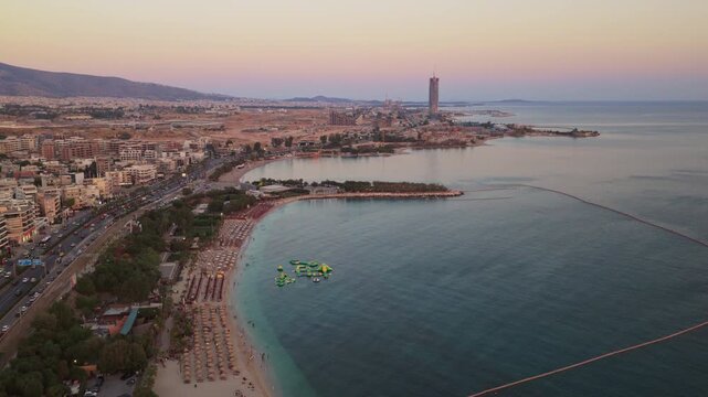 Stunning aerial footage showing the beautiful coastline of the Athens Riviera, Greece, featuring a sandy beach with umbrellas, a water park, and urban development during a colorful sunset