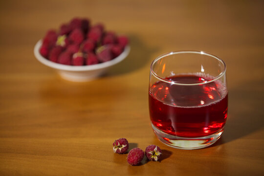 Glass of fresh red raspberry juice on a wooden table with blurred berries background.