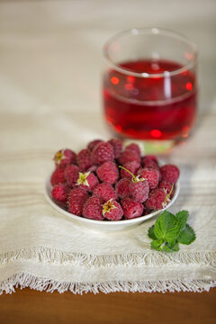 Fresh ripe red raspberries in a bowl with a blurred glass of juice on a tablecloth.