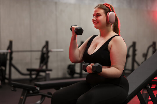Happy overweight woman with headphones exercising with dumbbells in gym, space for text
