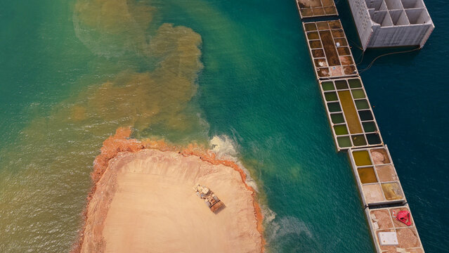 Land reclamation project advancing in the ocean. With heavy machinery forming an artificial island and creating visible silt and sediment plumes in the turquoise water