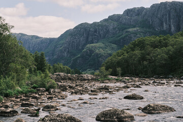 Majestic rocky mountains tower over a wide, stone-filled river flowing through a lush green valley. A serene and powerful natural landscape under a cloudy sky, perfect for outdoor adventure themes. © KatyaPulka