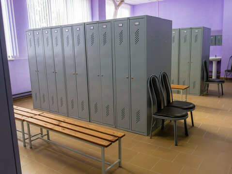Rows of tall grey metal storage lockers stand in a clean dressing room with wooden benches, black chairs, and purple walls.