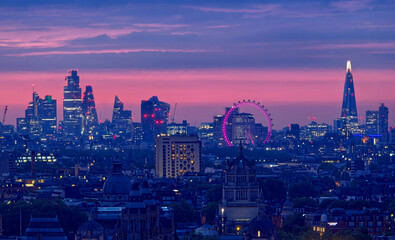 Panoramic view of the London skyline during the early morning blue hour, featuring the illuminated London Eye, The Shard, modern skyscrapers, historic architecture, and glowing city streets beneath a  © phillips