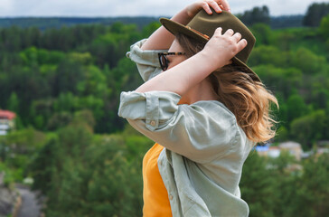 Profile portrait of stylish woman holding fedora hat on mountain viewpoint overlooking green forest valley. Idea of wanderlust, freedom and discovering nature. © 5ph