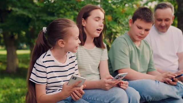 kids and adult on smartphones outdoors, children online while sitting in nature, preteens engaged with mobile devices outside, the digital generation relaxes outside with smartphones in hand