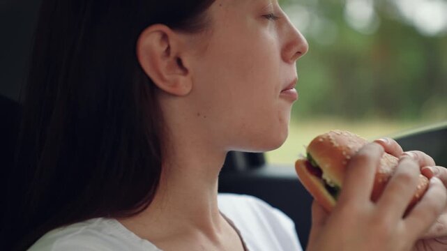 woman girl eating burger car, happy person, Girl eats in car, Tasty burger meal, Surrounded by trees, Golden bun, Hands close up, Quiet interior, Fresh ingredients, Concentrated expression, Peaceful