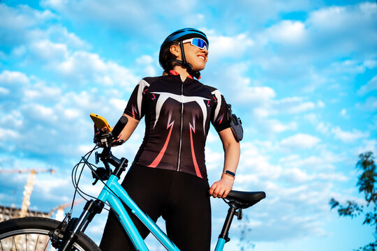 Low angle view of woman cyclist holding smartphone against blue sky. Modern sports, outdoor activity concept, mobile connection.