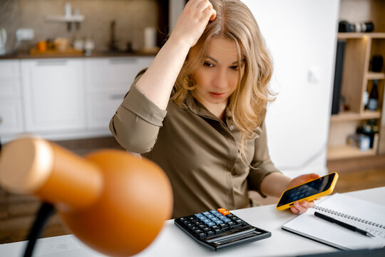 Confused woman calculating personal budget with calculator and smartphone at desk. Financial problem, inflation crisis concept, debt anxiety.