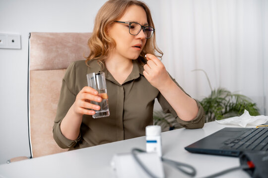 Sick woman taking pill with glass of water at office desk. Workplace healthcare, disease symptom concept, medication treatment.