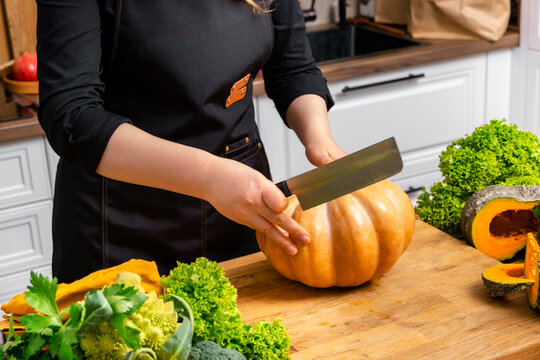 Woman chef cutting big ripe pumpkin with knife on wooden board. Autumn home cooking, raw organic pumpkin recipe, seasonal comfort food, healthy diet preparation.