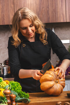 Woman chef carving large autumn pumpkin with knife in home kitchen. Seasonal pumpkin soup prep, autumn home cooking, organic healthy food concept, culinary autumn recipes.