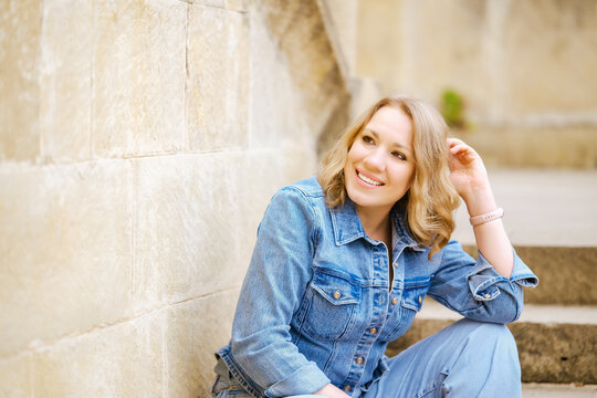 Smiling fair haired woman sitting on stone stairs outdoors. Concept of urban travel, city exploration and relaxation.