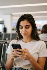 Fototapeta premium Young woman using a smartphone while waiting in an airport lounge, digital communication and travel technology