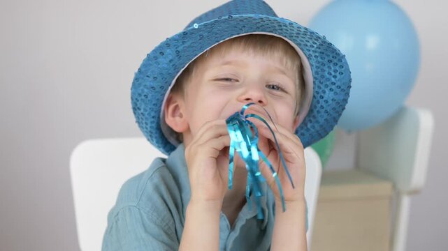 Boy in blue straw hat blows party noisemaker with big happy smile in front of colorful balloons. Joyful child birthday celebration and festive party concept. Family holiday, positive emotions and fun.