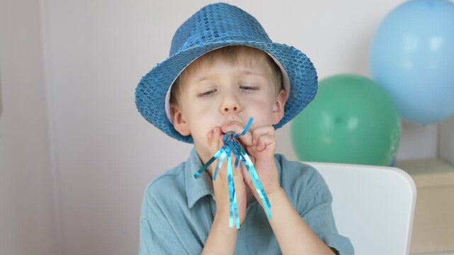 Smiling child celebrates birthday with party horn and decorations indoors. Cheerful emotions, special occasion, family holiday and joyful childhood celebration concept. Boy holds shiny noisemaker. 