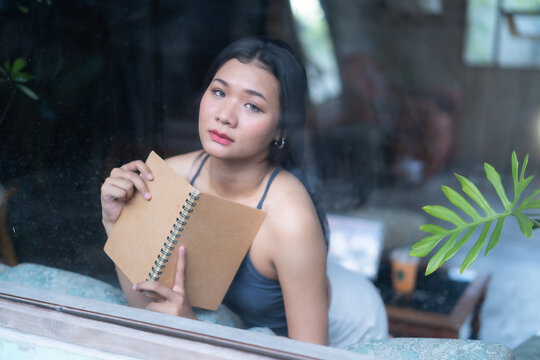 Asian woman sitting on sofa and looking out the window while reading a book, and gazed thoughtfully out of a large glass window in a cozy room with houseplants.