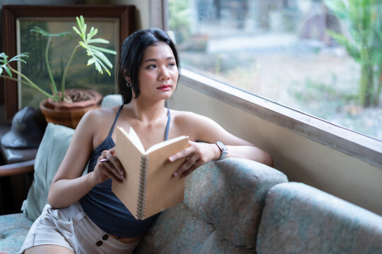 Asian woman sitting on sofa and looking out the window while reading a book, and gazed thoughtfully out of a large glass window in a cozy room with houseplants.