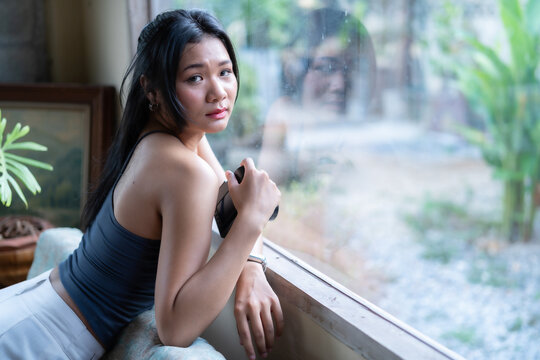 Young Asian female photographer holding a black mirrorless camera, looking thoughtfully out a large glass window in a cozy room with soft daylight.