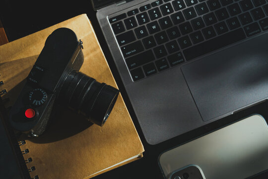 wooden table workspace setup featuring a laptop computer, a plastic cup of iced black coffee, modern smartphone, notebook, a pen, classic black rangefinder camera morning sunlight.