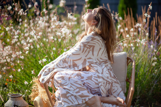 Young woman relaxes on chair in summer garden wearing stylish robe holding straw hat. Lady breathes, enjoys sun among flowers
