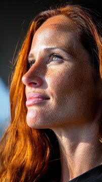 Young woman with long red hair and soft freckles gazes into the horizon while standing outside in the natural warm light of a peaceful afternoon in a quiet setting