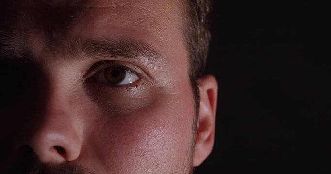 close-up of half of a man's face with a neutral, emotionless expression against a dark background.