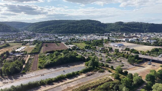 Drone aerial panorama of Trier, Germany, featuring hillside vineyards, the Moselle River, riverside woodland, a navigation lock and residential neighborhoods. Scenic summer landscape with rolling hill