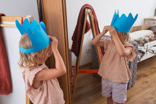  girl holding a handmade blue paper crown while looking at her reflection in a mirror.