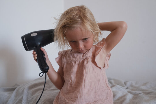 Funny cute child looking focused and independent while blowing dry her bedhead hair at home.