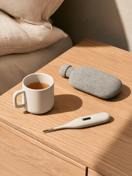 Tea mug digital thermometer hot water bottle wooden nightstand flu care still life minimalist sunlight shadow cozy
