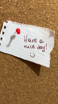 A hand pinning a handwritten note with the message Have a nice day and a smiling face onto a cork board with a red pushpin. The concept of positive wishes and vibes.