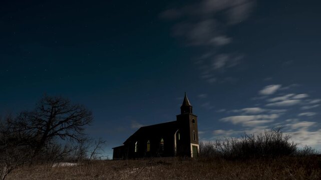 Time-lapse of an old, abandoned wooden church stands silhouetted against a starry night sky with moving clouds and bare trees.
