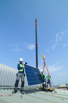 A team of solar farm laborers in green shirts and yellow hard hats calmly stream across the roof with panels held high, seen ultra wide high on an industrial warehouse roof in a Thai city.