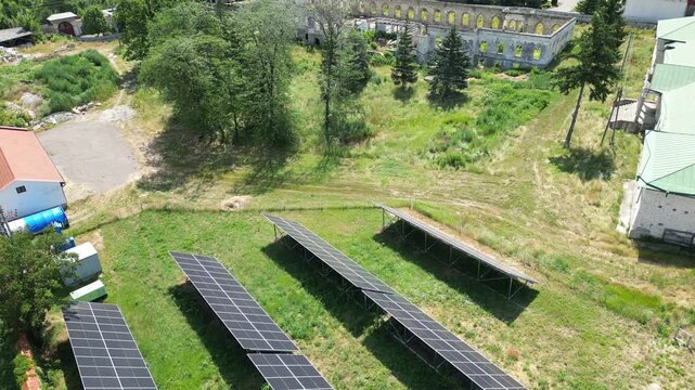 Aerial view of solar panels arranged in rows across a grassy field. Tall trees and green shrubs surround the installation. Large building with a dome appears in the distance