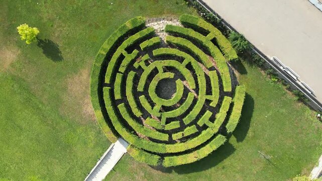 Circular hedge maze of neatly trimmed green hedges forms concentric patterns. Its central opening reveals a dark interior space. Grassy areas and pathways frame the maze's perimeter