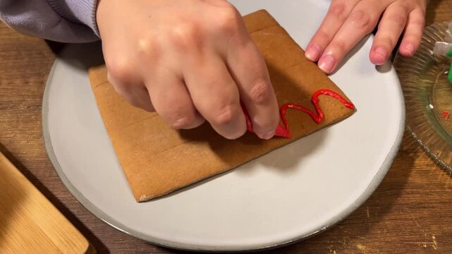 4K close up top view video. Female hand draws red wave pattern on gingerbread house roof in plate on wooden table. Woman with plastic bag with edible paint. Concept of sweet food, New Year 2027