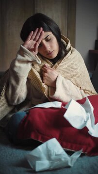 Young Hispanic woman sitting on her sofa at home with fever and chills. Health lifestyle video representing flu symptoms, illness, recovery, self-care and resting at home.
