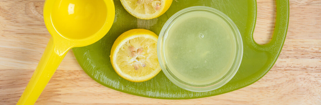 Squeezing real lemon juice, green cutting board, glass prep bowl, yellow citrus squeezer and fresh lemons cut in half on a wood countertop, preparation for baking at home
