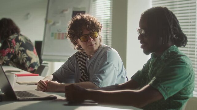 Gen Z Caucasian woman with yellow glasses discussing business data on laptop with Black male coworker in open space office filled with sunlight