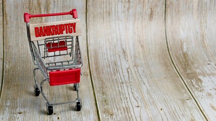 An empty shopping cart with a wooden block reading BANKRUPTCY on a textured background. © FaizAhmad