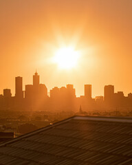 Obraz premium City skyline at sunset with an orange sky and sun shining brightly above urban buildings and rooftops.